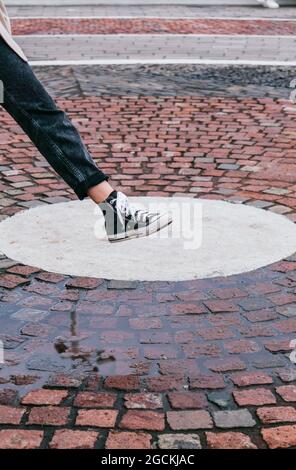 Unrecognizable Woman in stylish jeans and sneakers stepping in puddle on pavement while playing on street on rainy day Stock Photo