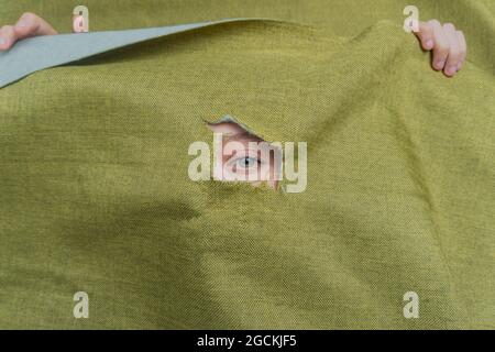 Unrecognizable young green eyed female peeking out through ripped hole in green cloth Stock Photo