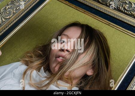 Top view of young long haired female with green eyes dressed in white blouse looking at camera through ornamental picture frame Stock Photo