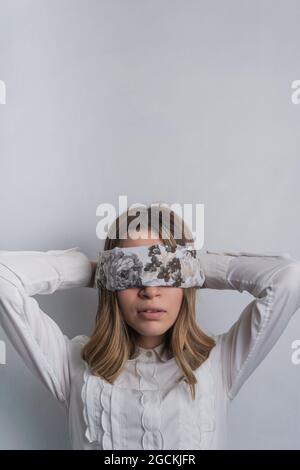 Young fair haired female in elegant blouse covering eyes with folded silk scarf with floral ornament against white background Stock Photo