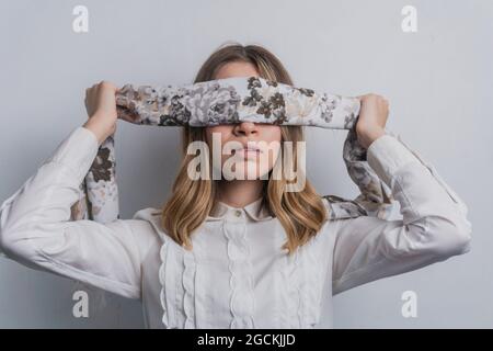 Young fair haired female in elegant blouse covering eyes with folded silk scarf with floral ornament against white background Stock Photo