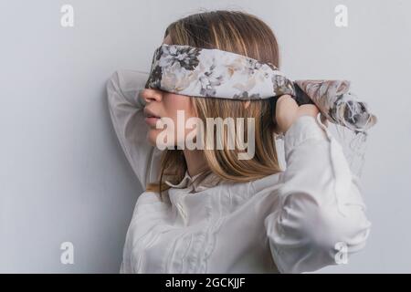Side view of young fair haired female in elegant blouse covering eyes with folded silk scarf with floral ornament against white background Stock Photo