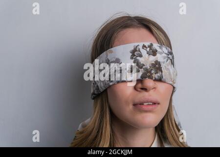 Young fair haired female in elegant blouse covering eyes with folded silk scarf with floral ornament against white background Stock Photo
