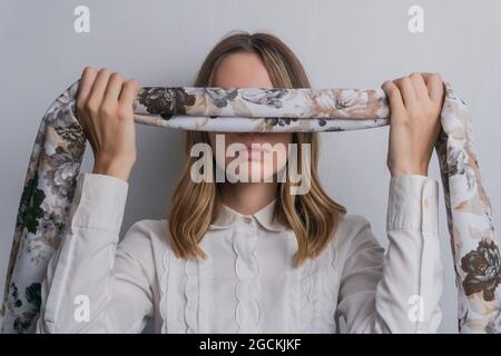 Young fair haired female in elegant blouse covering eyes with folded silk scarf with floral ornament against white background Stock Photo