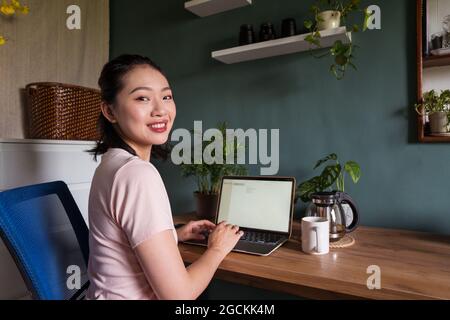 Side view of happy Asian female freelancer reading documents on laptop while sitting at table during remote work looking at camera Stock Photo