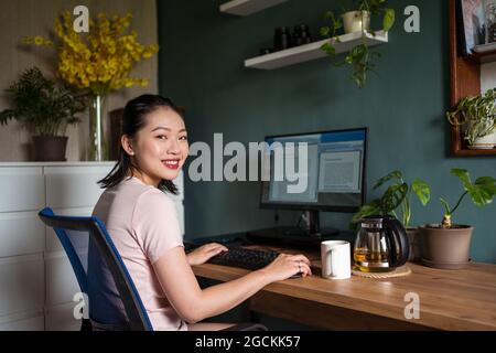 Side view of happy Asian female freelancer reading documents on computer monitor while sitting looking at camera at table during remote work Stock Photo