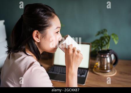 Side view of Asian female freelancer reading documents on laptop while sitting and drinking tea looking away at table during remote work Stock Photo