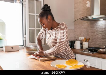 Young African American female crushing fresh plantain on cutting board while preparing patacones at home Stock Photo