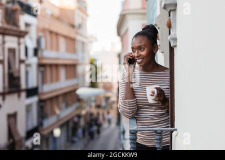African American female with cup of hot drink talking cellphone on balcony Stock Photo