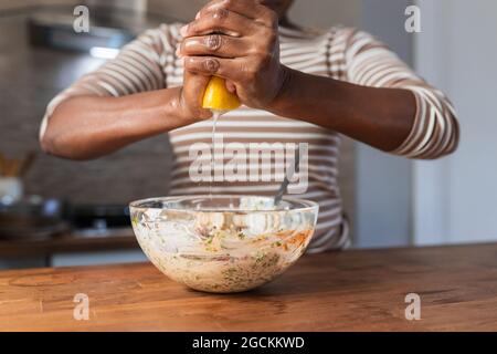 Crop unrecognizable ethnic female squeezing fresh lemon over bowl with food at table in kitchen Stock Photo
