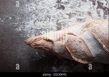 Appetizing freshly baked baguette with crispy crust placed on wooden table covered with white flour against black background Stock Photo