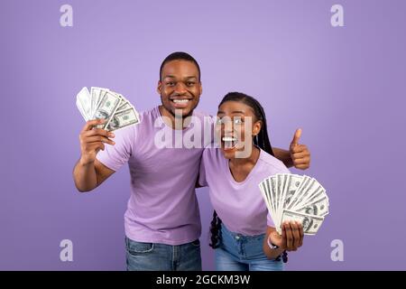 Portrait of overjoyed young African American couple holding and showing bunch of money, celebrating success together, making thumb up gesture, posing Stock Photo