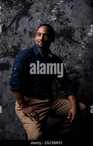 Serious stylish ethnic male sitting on wooden stool in studio on dark background and looking away Stock Photo