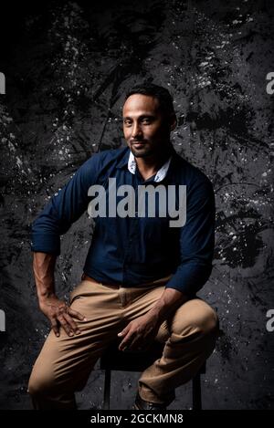Serious stylish ethnic male sitting on wooden stool in studio on dark background and looking at camera Stock Photo