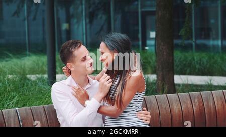 Young multiethnic adults stroking each other and talking on date outdoors. Black woman sitting on man's knees. Stock Photo