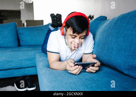 Delighted focus Latin teen boy in headphones playing videogame on mobile phone while lying on sofa at home Stock Photo