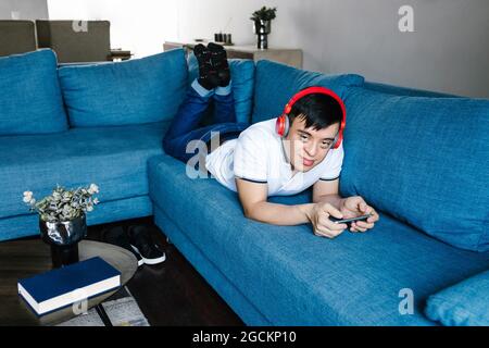 Delighted Latin teen boy in headphones playing videogame on mobile phone while lying on sofa at home and looking at camera Stock Photo
