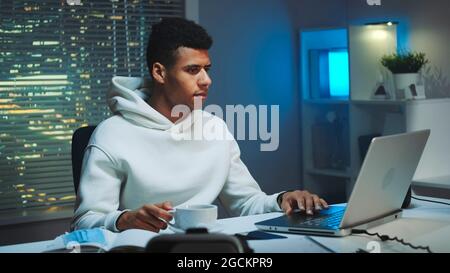 Handsome multiracial man drinking a cup of coffee and working on computer at night. There are skyscrapers in the background. Stock Photo