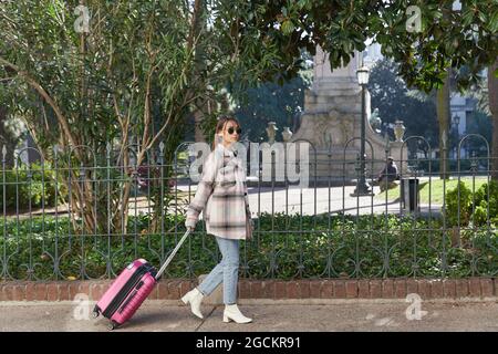Full body side view of young female traveler in stylish outfit and sunglasses pulling pink suitcase while walking on pavement in city Stock Photo
