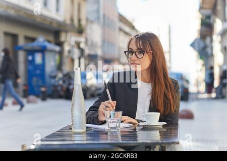 Positive young business lady in elegant suit and eyeglasses taking notes in notebook while sitting at table in outdoor cafe in city looking away Stock Photo