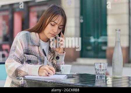 Young Woman taking notes in notebook during phone conversation while sitting at table in outdoor cafe in city Stock Photo