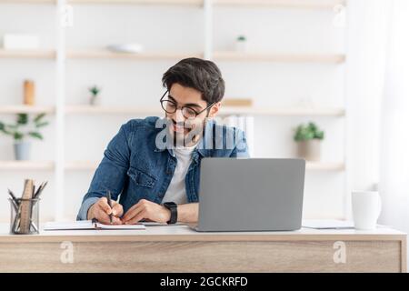Positive arab male manager working from home during COVID-19 pandemic, using modern laptop and taking notes Stock Photo