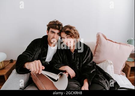 Content couple in bathrobes relaxing on couch at home while hugging and looking at camera Stock Photo