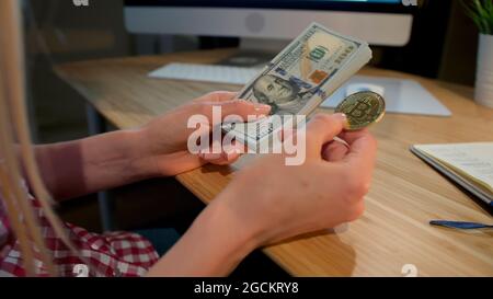 Crop view of female hands holding metal bitcoin and bundle of dollar banknotes sitting at wooden desk with notepad and computer monitor at night Stock Photo