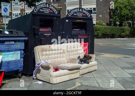 Old sofa and clothes dumped in the street by bins, Portsmouth City, Hampshire, UK Stock Photo