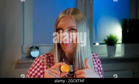 Cheerful young blond woman in casual clothes sitting at dark window at night holding bitcoin and doing thumbs up gesture smiling and looking at camera Stock Photo