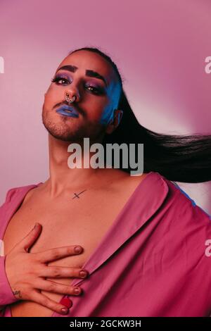 Portrait of glamorous transgender bearded Woman in sophisticated make up posing against pink background at studio looking at camera Stock Photo