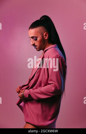 Portrait of glamorous transgender bearded Woman in sophisticated make up putting on shirt against pink background at studio Stock Photo