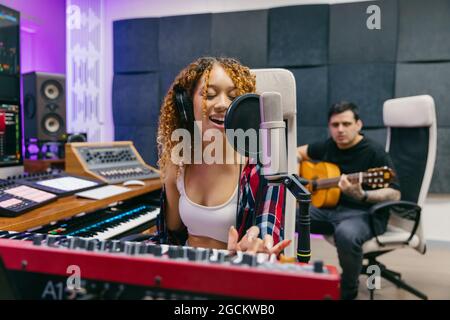 Happy male guitarist playing acoustic guitar against black female partner in headphones singing into microphone in recording studio Stock Photo