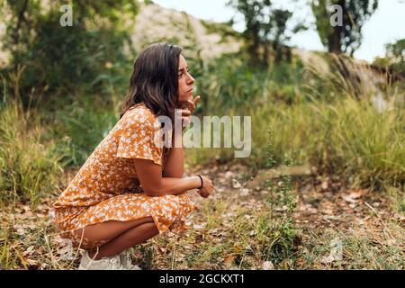 Thoughtful beautiful brunette woman wearing a dress squatting in the woods Stock Photo
