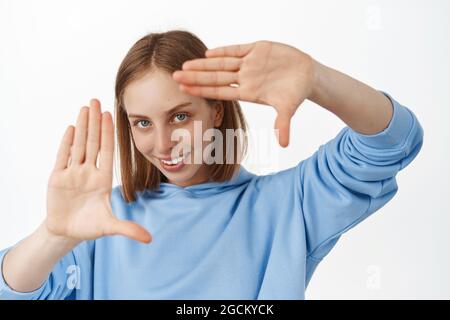 Image of creative young woman looking through hand frames gesture with pleased thoughtful smile, creating perfect shot, searching right angle Stock Photo