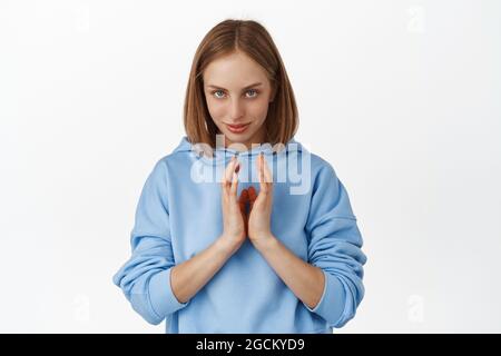Image of cunning girl, smiling sly and steeple fingers like evil genius, has plan, thinking about something devious, scheming, standing against white Stock Photo