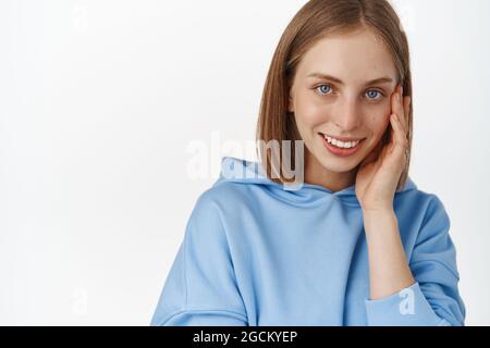 Women face and skin care. Portrait of beautiful blond girl touching her cheek, smiling and gazing happy at camera, dont be shy drop your cosmetics Stock Photo