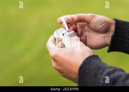 Crop hands of unrecognizable male with true wireless earphones in white case on background of blurred green grass Stock Photo