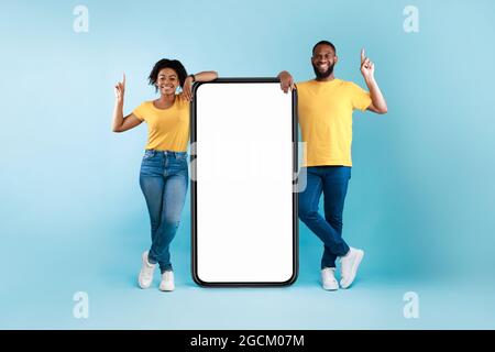 Cheerful Afro couple standing next to big smarthone with blank screen, pointing upwards, recommending mobile app, mockup Stock Photo
