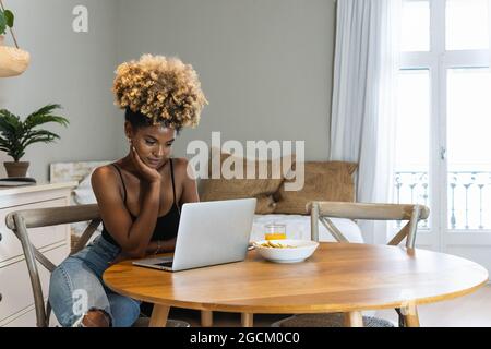 Attractive African American female freelancer sitting on chair and watching laptop at table with breakfast Stock Photo