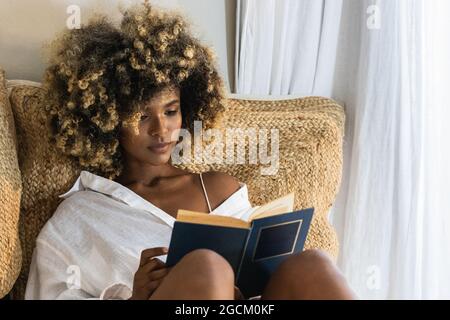 Attractive African American female with curly hair sitting on unmade bed and reading book in morning Stock Photo