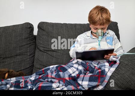 Sick boy in oxygen mask sitting on sofa at home and playing video game on tablet Stock Photo
