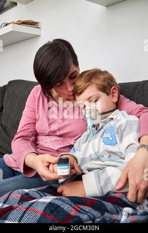 Caring Woman using pulse oximeter on finger of ill boy in oxygen mask using nebulizer during inhalation at home Stock Photo