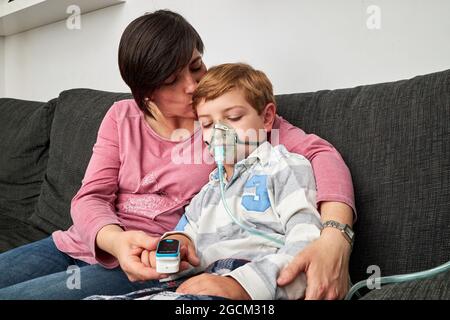 Caring Woman using pulse oximeter on finger of ill boy in oxygen mask using nebulizer during inhalation at home Stock Photo