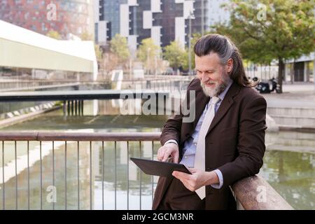 Positive respectable middle aged bearded man in formal outfit browsing tablet while standing on embankment in city Stock Photo