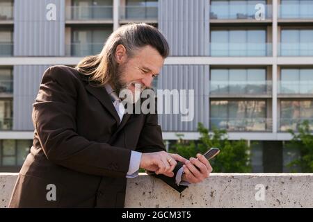 Side view of smiling bearded mature male with gray hair wearing classy suit browsing on smartphone while standing against modern city buildings Stock Photo