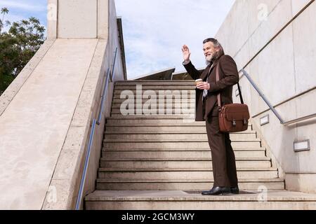 Low angle full body side view of stylish mature bearded male in elegant suit with briefcase and takeaway coffee waving hand and looking away while sta Stock Photo