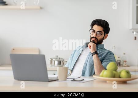 Thoughtful arab freelancer man looking at laptop screen while working remotely at home, sitting at kitchen with computer Stock Photo