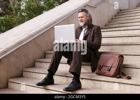 Positive middle aged bearded male entrepreneur in formal clothes sitting on stairway and working online on laptop in city Stock Photo