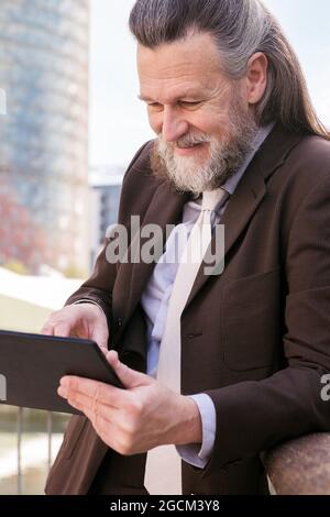 Positive respectable middle aged bearded man in formal outfit browsing tablet while standing on embankment in city Stock Photo